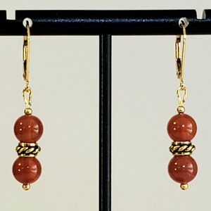 May include: A pair of gold-toned leverback earrings, each featuring two smooth, round, reddish-brown beads separated by a small, textured, antique-gold-colored metal spacer bead. The earrings hang vertically from simple gold-toned metal hooks.