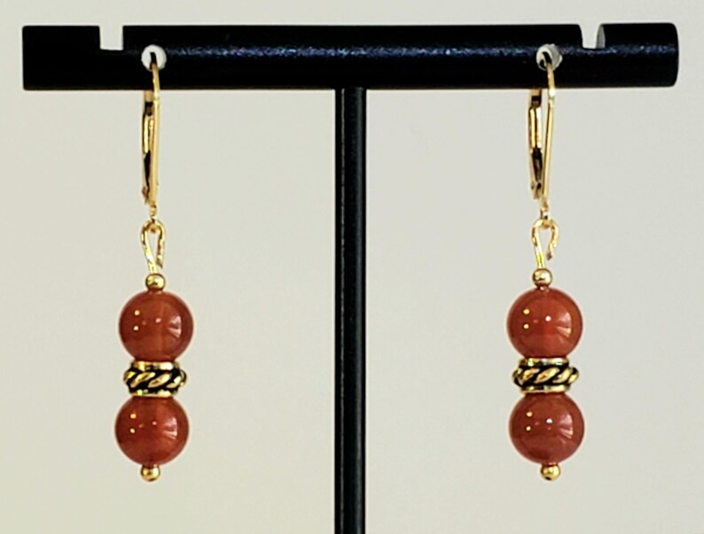 May include: A pair of gold-toned leverback earrings, each featuring two smooth, round, reddish-brown beads separated by a small, textured, antique-gold-colored metal spacer bead. The earrings hang vertically from simple gold-toned metal hooks.