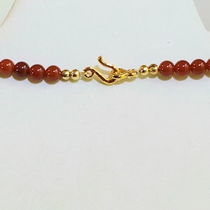 May include: A necklace featuring a series of round, deep red carnelian beads interspersed with small, polished gold-toned metal spheres. The piece is secured with a gold-toned metal hook-and-eye clasp centered between the bead strands.