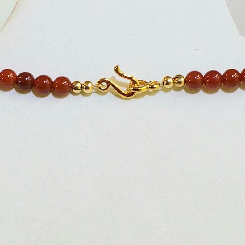 May include: A necklace featuring a series of round, deep red carnelian beads interspersed with small, polished gold-toned metal spheres. The piece is secured with a gold-toned metal hook-and-eye clasp centered between the bead strands.