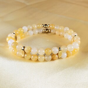 May include: A double-strand bracelet featuring round, yellow and white beads. Gold-colored metal accents are placed between the strands and at the clasp. The bracelet is displayed on a yellow satin surface.