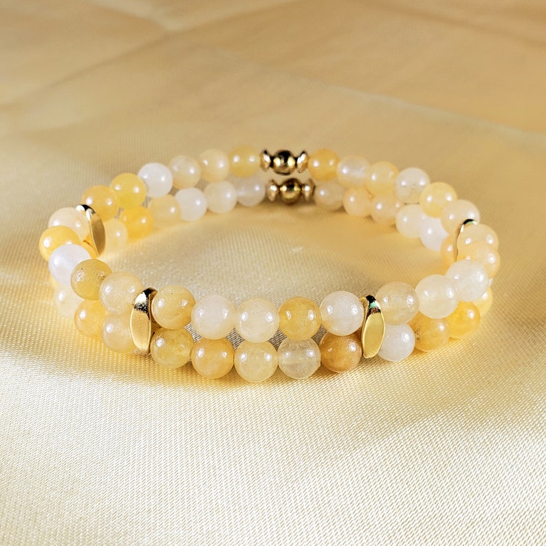May include: A double-strand bracelet featuring round, yellow and white beads. Gold-colored metal accents are placed between the strands and at the clasp. The bracelet is displayed on a yellow satin surface.