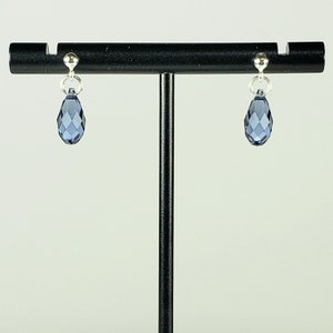 May include: A pair of silver-toned drop earrings with faceted, teardrop-shaped blue crystals. The earrings are suspended from small silver studs and are displayed on a black jewelry stand. The crystals have a multi-faceted design.