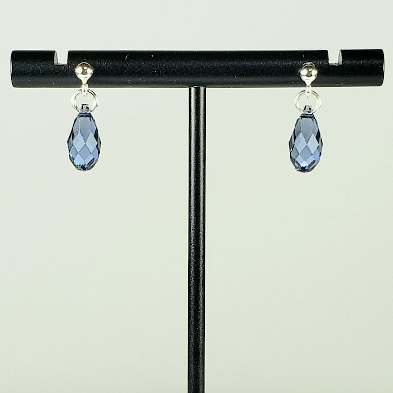 May include: A pair of silver-toned drop earrings with faceted, teardrop-shaped blue crystals. The earrings are suspended from small silver studs and are displayed on a black jewelry stand. The crystals have a multi-faceted design.