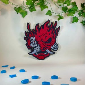 Op de afbeelding: Een rood en zwart embleem met een gestileerd oni-maskerontwerp met vlammen. Het masker heeft een zilver en wit ontwerp aan de linkerkant. Blauwe, ovaalvormige stenen zijn eronder verspreid.