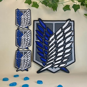 May include: Patches featuring the Survey Corps emblem from the anime Attack on Titan. The patches show a blue and white wing design on a grey background. Several blue, oval-shaped embellishments are scattered below the patches.