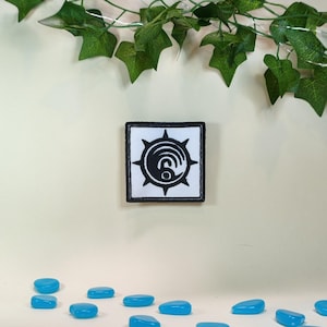 Puede incluir: Parche cuadrado con un diseño en blanco y negro. El parche presenta un diseño central en espiral rodeado por un borde puntiagudo. El parche está bordeado con costuras negras. El fondo es de color beige claro con piedras azules.