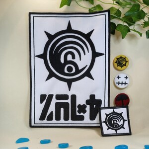 May include: A rectangular white fabric patch featuring a black embroidered sun-like emblem with a central spiral and stylized black text below. A smaller square patch with the same emblem, plus three circular pins with distinct graphic designs.