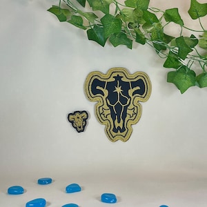 Peut inclure: Deux écussons brodés avec un motif noir et or. Le plus grand écusson présente un emblème détaillé, tandis que le plus petit reprend le motif. Du feuillage vert et des pierres bleues sont en arrière-plan.