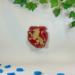 Puede incluir: Un parche bordado en rojo y dorado con un león dorado rampante sobre un escudo. El parche tiene un borde dorado y está sobre un fondo blanco con follaje verde.