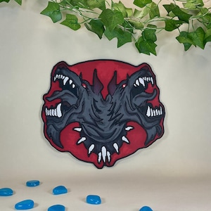 Op de afbeelding: Een gestileerde illustratie van een driekoppige hond, weergegeven in grijstinten en zwart, tegen een rode achtergrond. De hond heeft rode ogen en scherpe tanden. Het kunstwerk is waarschijnlijk een decoratief item.