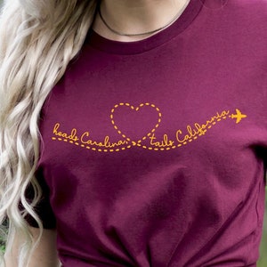 Könnte beinhalten: Ein maroonfarbenes T-Shirt mit einer gelben Grafik eines Herzens, durch das ein Flugzeug fliegt. Der Text "Head Carolina, Tail California" ist entlang der Flugbahn des Flugzeugs geschrieben.