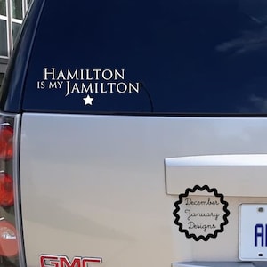May include: A white vinyl sticker on the back of a silver SUV. The sticker says "HAMILTON IS MY JAMILTON" with a white star. Below the sticker is a black and white sticker with the text "December January Designs".