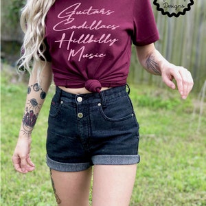 Puede incluir: Una mujer lleva una camiseta granate con el texto "Guitars & Cadillacs Hillbilly Music" en letras rosas. Lleva pantalones cortos de mezclilla negros y tiene tatuajes en los brazos.