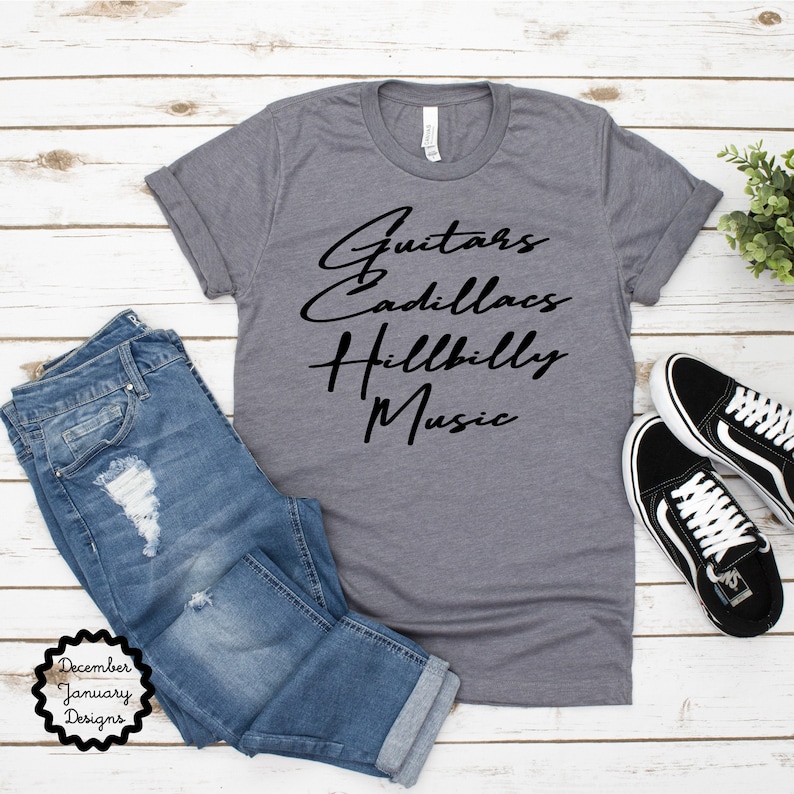 Puede incluir: Una camiseta gris jaspeada con texto negro que dice "Guitars Cadillacs Hillbilly Music". La camiseta est&aacute; puesta plana sobre una superficie de madera blanca con un par de pantalones vaqueros azules y zapatillas negras a su lado.