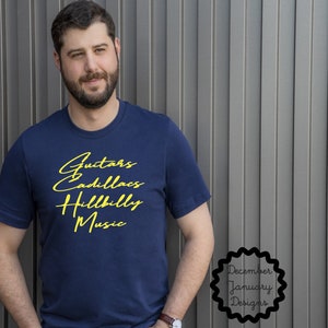 Puede incluir: Una camiseta azul marino con texto amarillo que dice "Guitars Cadillacs Hillbilly Music". El texto est&aacute; en una fuente de letra manuscrita. La camiseta la lleva puesta una persona de pie frente a una pared gris. Un peque&ntilde;o logotipo en blanco y negro en la esquina inferior derecha dice "December January Designs".