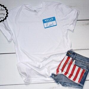 May include: White t-shirt with a blue name tag that says "Hello, my name is Earl" on it. The t-shirt is laying on a white wooden surface with a pair of denim shorts with red and white stripes on the back pocket.