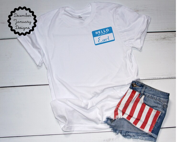 May include: White t-shirt with a blue name tag that says "Hello, my name is Earl" on it. The t-shirt is laying on a white wooden surface with a pair of denim shorts with red and white stripes on the back pocket.