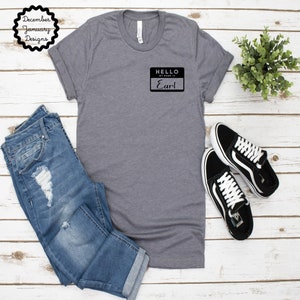 May include: A gray t-shirt with a black name tag that says "Hello, My Name Is Earl" on a white wooden background.
