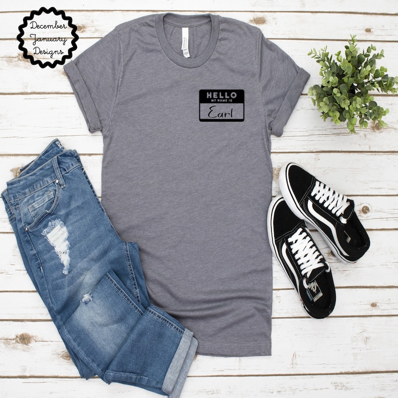 May include: A gray t-shirt with a black name tag that says "Hello, My Name Is Earl" on a white wooden background.