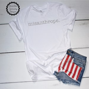 May include: White t-shirt with the text "misanthrope." printed in gray on the front. The t-shirt is laying on a white wooden surface with a pair of denim shorts with red and white stripes on the bottom.