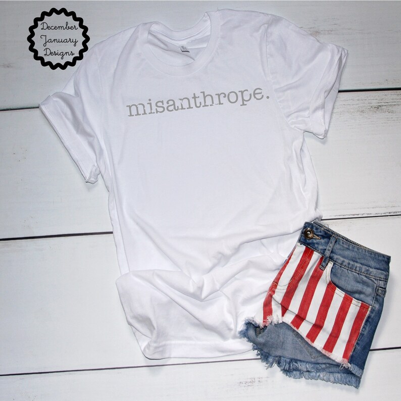 May include: White t-shirt with the text "misanthrope." printed in gray on the front. The t-shirt is laying on a white wooden surface with a pair of denim shorts with red and white stripes on the bottom.