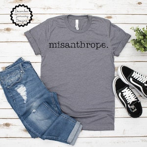 May include: Gray t-shirt with the word "misanthrope" printed in black on the front. The shirt is shown on a model wearing blue jeans and black sneakers.