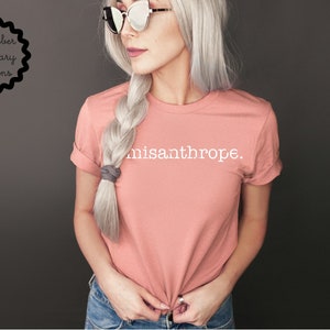 May include: A woman wearing a light pink t-shirt with the text "misanthrope." printed in white. She has long, silver hair braided and tied with a gray ribbon. She is wearing blue jeans and sunglasses.