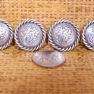 May include: Four silver-toned metal buttons with a rope-like border and an engraved floral design. The buttons are arranged in a row on a woven brown surface.