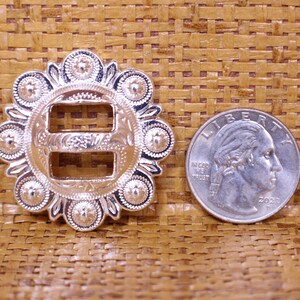 May include: Silver-toned metal buckle with a decorative design and two slots. The buckle is about the size of a US quarter.