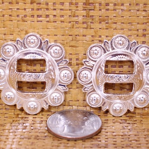 May include: Two silver-toned metal buckles with a decorative design. Each buckle has a circular shape with a raised edge and a center piece with a double-loop design. The buckles are surrounded by a ring of small, raised circles.