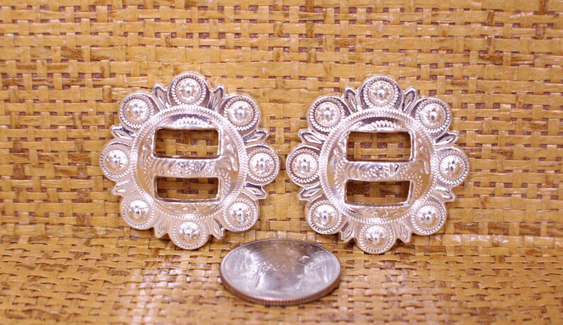 May include: Two silver-toned metal buckles with a decorative design. Each buckle has a circular shape with a raised edge and a center piece with a double-loop design. The buckles are surrounded by a ring of small, raised circles.