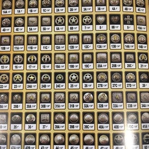May include: A collection of silver and gold metal belt buckles with various designs, including stars, crosses, hearts, and horseshoes. The buckles are arranged in rows and columns on a yellow background. Each buckle has a letter and number designation.
