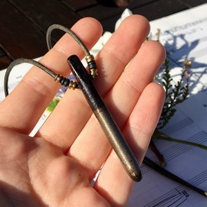 May include: A black and gold metal pendant necklace with a leather cord. The pendant is a long, thin, cylindrical shape with a textured surface.