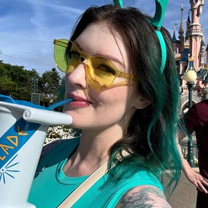 May include: A person wearing yellow-tinted sunglasses and a teal headband with pointed accents. They are holding a large white cup with the word "LAD" visible. The person is wearing a teal tank top and has teal-colored hair.