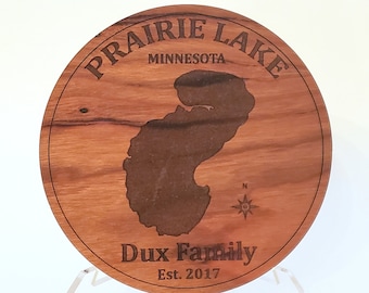 Puede incluir: Una placa redonda de madera con el texto "Prairie Lake Minnesota" y "Dux Family Est. 2017" grabado en ella. La placa presenta un mapa del lago y una brújula.