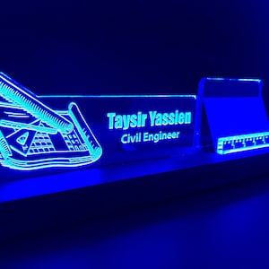 May include: A blue illuminated desk nameplate with a drawing of a compass, ruler, and pencils. The nameplate reads "Taysir Yassien, Civil Engineer".