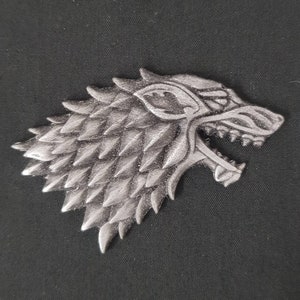 May include: A silver-colored metal wolf head with a detailed design. The wolf's fur is depicted with a textured, scaly pattern. The wolf's mouth is open, showing sharp teeth.