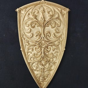 May include: A gold-colored, shield-shaped decorative item with intricate, swirling floral patterns. The design is raised, creating a three-dimensional effect. The shield has a pointed base and a decorative top element.