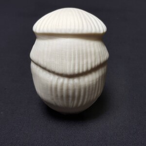 May include: A white ceramic egg-shaped object with a textured surface. The object has a rounded top and a slightly pointed bottom.