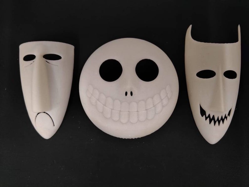 May include: Three white Halloween masks with different facial expressions. The masks are made of a smooth, white material and have black details. The first mask has a sad expression, the second mask has a wide smile, and the third mask has a sinister grin.