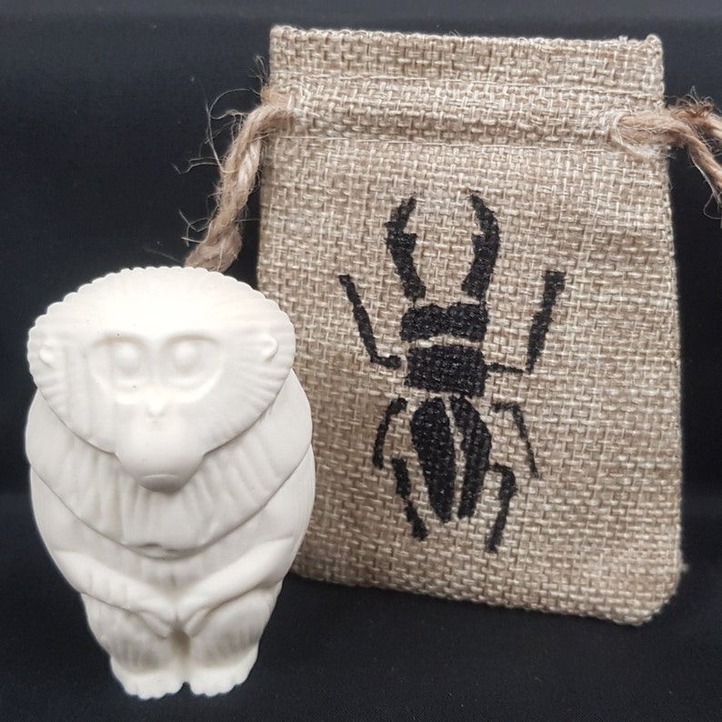 May include: White ceramic monkey figurine with a detailed face and sitting pose. The monkey is in front of a small burlap bag with a black beetle design.