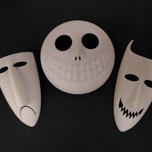 May include: Three white masks with black details. The masks are shaped like faces with different expressions. The mask on the left has a sad expression, the mask in the middle has a wide smile, and the mask on the right has a mischievous grin.