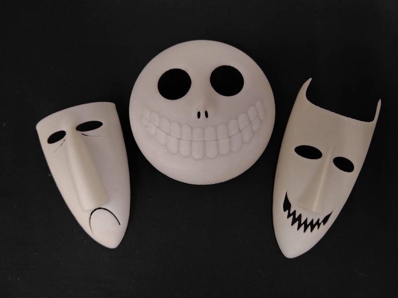 May include: Three white masks with black details. The masks are shaped like faces with different expressions. The mask on the left has a sad expression, the mask in the middle has a wide smile, and the mask on the right has a mischievous grin.