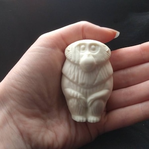 May include: White ceramic figurine of a monkey sitting with its hands clasped in front of it. The monkey has a detailed face with large eyes and a small mouth.