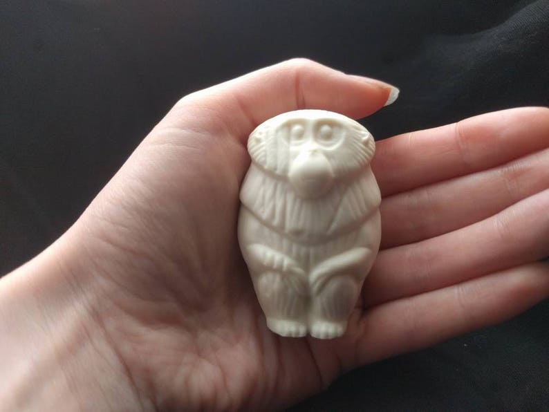 May include: White ceramic figurine of a monkey sitting with its hands clasped in front of it. The monkey has a detailed face with large eyes and a small mouth.