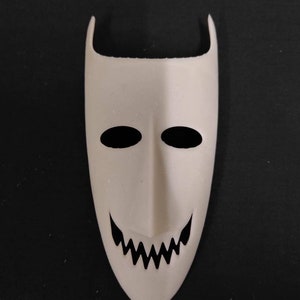 May include: White plastic mask with black eyes and a jagged smile. The mask has a pointed top and a rounded bottom.