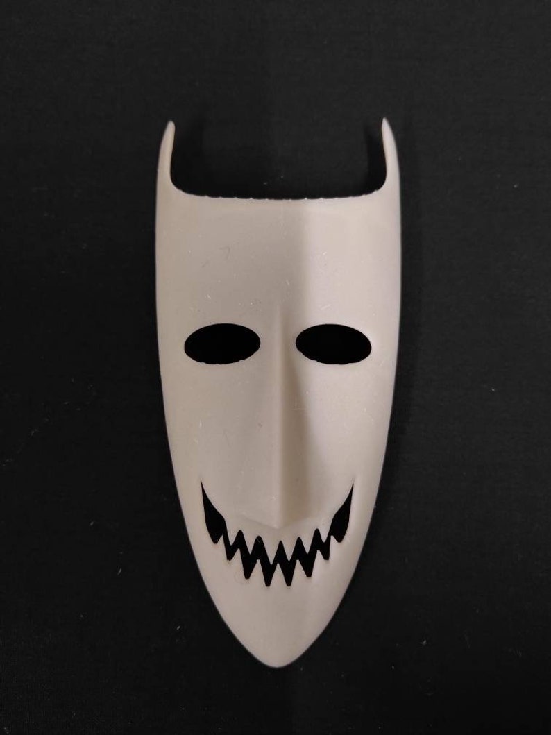 May include: White plastic mask with black eyes and a jagged smile. The mask has a pointed top and a rounded bottom.
