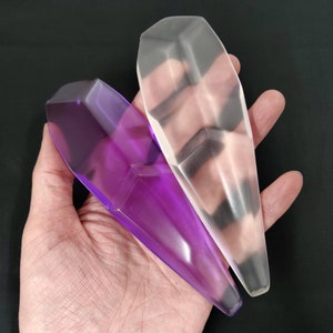 May include: Two faceted, elongated crystal-shaped objects. One is a translucent purple, the other is clear. The objects have a geometric design with multiple flat surfaces. The crystals are held in a hand against a black background.