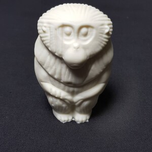 May include: A white ceramic figurine of a monkey sitting with its hands clasped in front of it. The monkey has a detailed face with large eyes and a small nose.